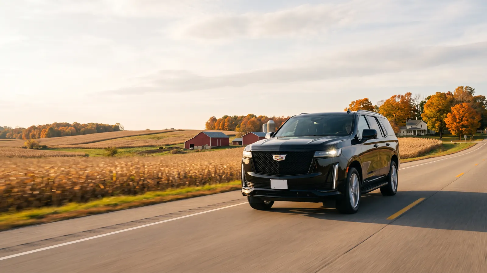 A black Escalade drives along a two-lane country highway past red barns and autumn trees under a wide sky